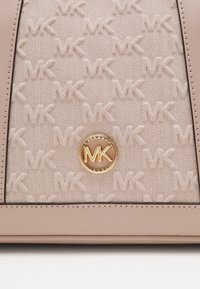 Beżowa, teksturowana tkanina z wytłoczonym wzorem "MK" oraz okrągłym złotym emblematem z literami "MK" i "Michael Kors", otoczonym gładkim skórzanym wykończeniem.