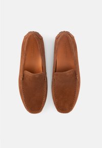 Ζευγάρι καφέ μοκασίνια από σουέτ με ραμμένα άκρα και επενδύσεις από δέρμα με λογότυπο, φωτογραφημένα από επάνω σε λευκό φόντο.