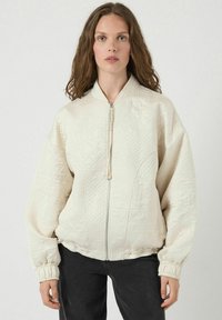 Giacca bomber crema trapuntata con colletto alto, chiusura con zip, polsini elastici e motivi testurizzati sulla stoffa.