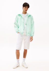 Muntgroene lichtgewicht jas met capuchon, wit t-shirt, witte shorts en witte sneakers, staand tegen een effen lichte achtergrond.
