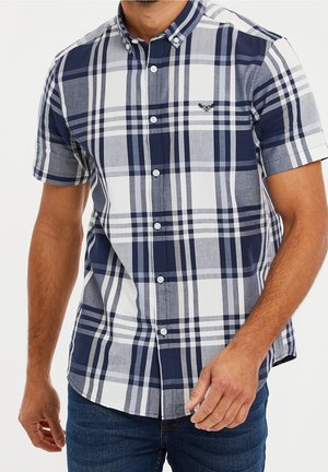 Mann trägt ein kurzärmliges Hemd mit Knopfleiste, kariert in Marineblau, Grau und Weiß, mit kleinem, auf der Brust gesticktem Logo.