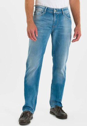 Homme portant un jean bleu clair, une chemise blanche et des chaussures en cuir marron foncé, debout devant un fond blanc uni.