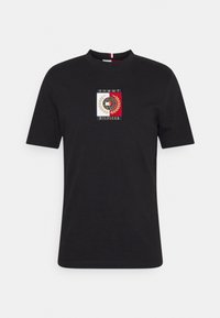 Černé tričko s krátkým rukávem vyrobené z bavlny. Obsahuje čtvercové logo se zlatou výšivkou a červené, bílé a modré obdélníkové prvky.
