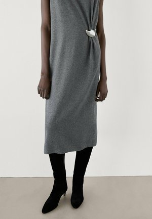 Abito grigio lavorato a maglia con un dettaglio laterale annodato che presenta un accento bianco. Indossato con stivali neri sopra il ginocchio, completato da una texture liscia.