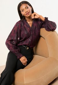 Burgundy sheer blouse with black floral pattern, puffed long sleeves, and a buttoned front. Worn with black trousers and a gold-buckled belt.
