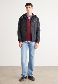 Sort letvægtsjakke med hætte, rød struktureret sweater, blå stribet skjorte, lyseblå jeans og mørke sneakers. Enkel, lagdelt outfit.