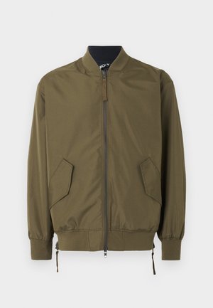 Νέος άνδρας που φοράει ένα χακί μπουφάν με φερμουάρ, λευκό T-shirt, ανοιχτό μπλε τζιν σορτς, λευκές κάλτσες και μαύρα μοκασίνια, στέκεται με το ένα χέρι στην τσέπη.