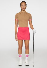 Bruine gestructureerde polo met roze accenten, gecombineerd met een roze golfrok. Witte sneakers en roze gestreepte sokken maken de outfit compleet.