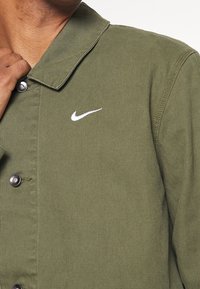 Vihreä, teksturoitu kangaspaita, jossa on kaulus ja napsautettavat napit. Vasemmassa rinta-alueessa on valkoinen Nike-logo.