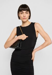 Moteris su trumpais tamsiais plaukais, vilkinti juodą bevėrę suknelę, ant peties nešiojanti juodą rankinę su auksine grandinėlės dirželiu.