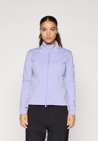 Žena s kučeravými vlasmi, oblečená v svetlom fialovom zip-up bundu a čiernych nohaviciach, stojí pred jednoduchým svetlým pozadím.