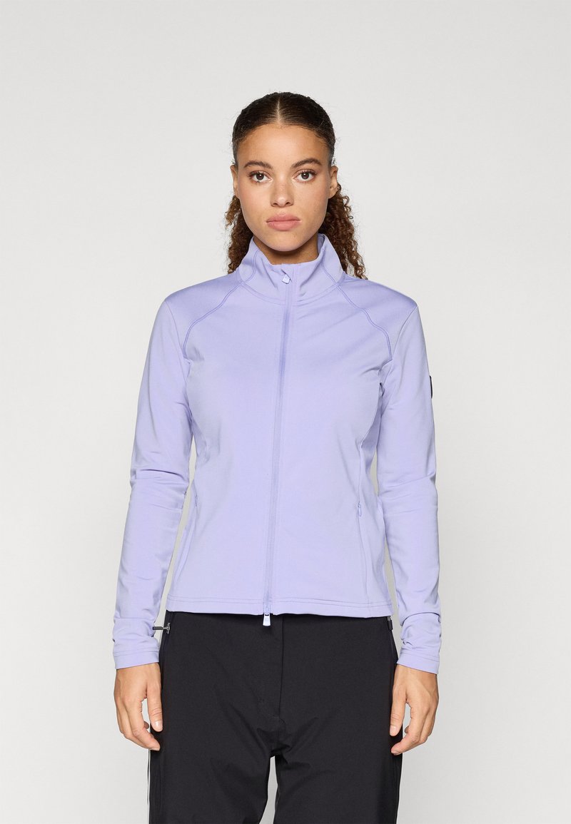 Žena s kučeravými vlasmi, oblečená v svetlom fialovom zip-up bundu a čiernych nohaviciach, stojí pred jednoduchým svetlým pozadím.