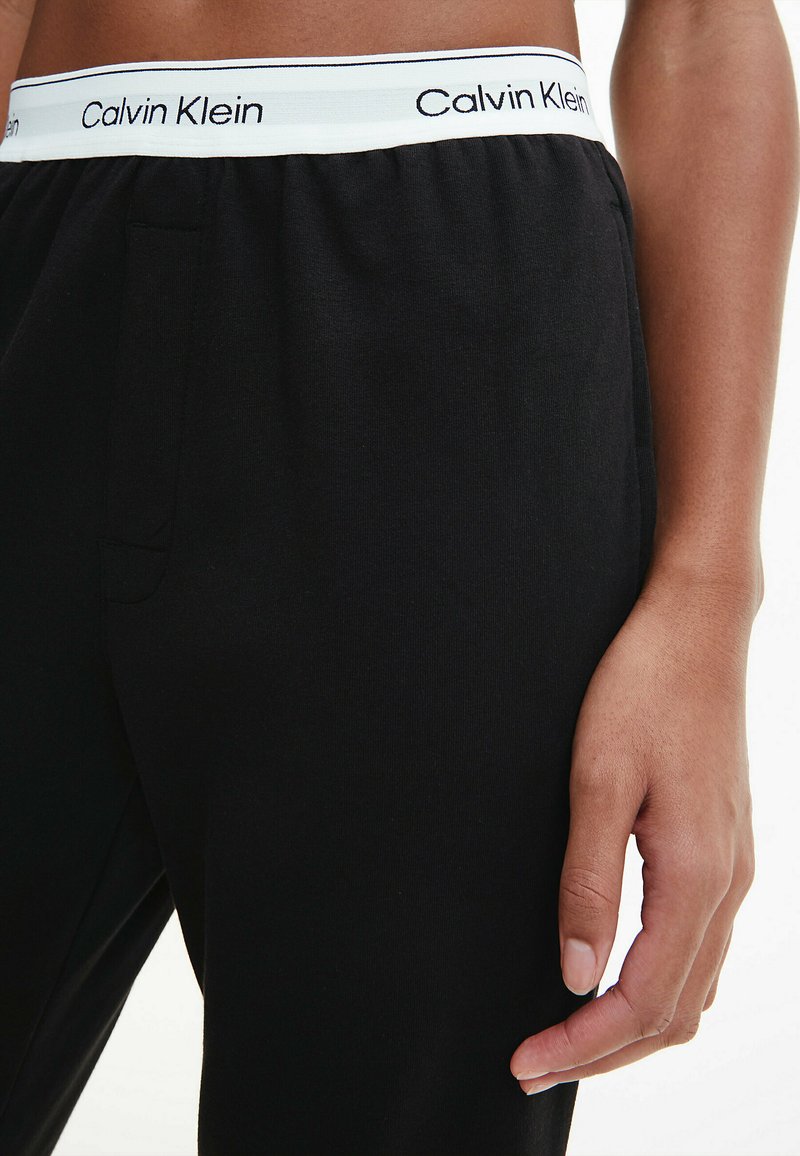Schwarze Baumwoll-Jogginghosen mit einem elastischen Bund, der ein weißes Calvin Klein-Logo und eine Seitentasche aufweist, und zeigen eine glatte Textur.