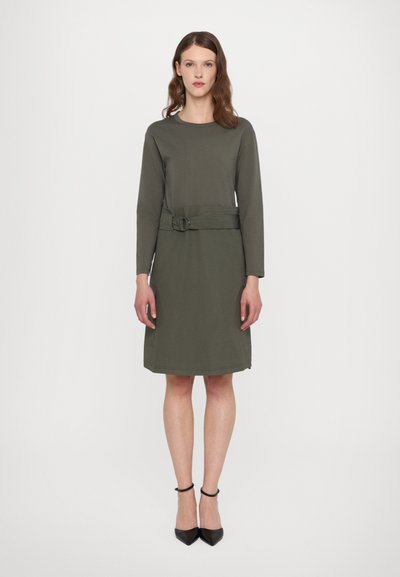 Olivengrünes Kleid mit langen Ärmeln und tailliertem Design mit metallischem Ringverschluss. Glattes Material und knielanger Schnitt. Schwarze hohe Absätze.