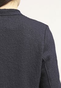 Pull bleu marine foncé avec un tissu texturé, doté d'un col simple et d'un design épuré. Le dos présente une coupe minimaliste sans coutures.