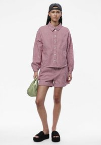 Giacca a righe rossa e bianca con bottoni e due tasche frontali, abbinata a pantaloni corti coordinati; tiene una borsa a quadretti verde; sandali neri con tacco alto.