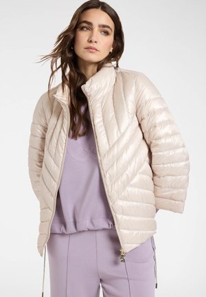 Kvinde i jakke i lysebeige quiltet stof over en lavendelfarvet sweatshirt og matchende bukser, stående foran en helt hvid baggrund.