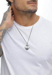 Sølv anker vedhængs halskæde med sorte detaljer, hængende fra en sølv kæde, båret over en enkel hvid t-shirt.