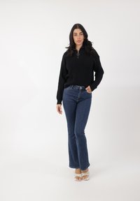 Suéter negro de punto con cuello con cremallera, combinado con jeans acampanados de cintura alta en azul oscuro. La modelo lleva sandalias de tacón blancas. Fondo blanco liso.