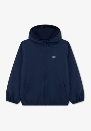 Giacca leggera blu navy con cappuccio, cerniera frontale, polsini elastici e tasche laterali. Presenta un piccolo logo Lacoste sul petto.