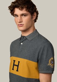 Sivá polo košeľa s žltým pruhom, ktorá má veľké vyšívané "H" a logo nášivku na rukáve. Vyrobená z jemnej bavlnenej zmesi.