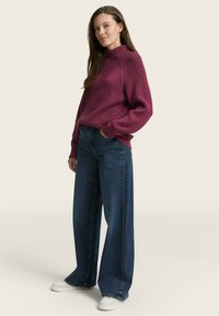Burgunderfarbener, gerippter Rollkragenpullover kombiniert mit weit geschnittenen, dunkelblauen Jeans und weißen Sneakers, was einen entspannten, lässigen Look kreiert.
