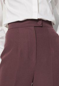 Personne portant un pantalon bordeaux taille haute, ajusté, avec un gros bouton et une chemise blanche rentrée à l'intérieur, vue de la taille au milieu de la cuisse.