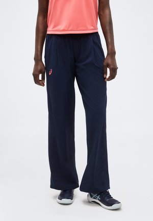 Άτομο που φοράει ναυτικό μπλε παντελόνια με φαρδιά μπατζάκια με ροζ λογότυπο, κοραλί αμάνικο τοπ και ναυτικά μπλε αθλητικά παπούτσια, στέκεται σε λευκό πάτωμα.