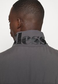 Grå jakke med høj krave, der har et sort, blankt logo på ryggen. Stoffet ser glat ud med en mat overflade.
