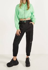 Mintgrön kort jacka med dragkedja fram och elastiska ärmar, kombinerad med svarta cargobyxor med fickor och elastiska fållar, bärs tillsammans med chunky svarta sneakers.