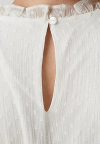 Blouse blanche avec de fines rayures verticales, des points texturés, un col à volants et un dos en forme de clé maintenu par un bouton.