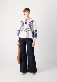 Weiße Bluse mit blauen Blumenstickereien, elastischen Bündchen und einer dekorativen Schleife. Dunkle, weit geschnittene Jeans. Braune Handtasche und spitze Schuhe.