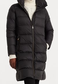 Svart puffjacka med en quiltad design, hel dragkedja och huva. Har ribbade ärmmuddar och sidofickor med dragkedja för funktionalitet.