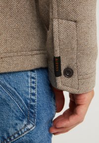 Brun herringbone-jacka med ärm som har en svart etikett med "CHASIN'" och en mörk knapp. Buren tillsammans med blå denimjeans.