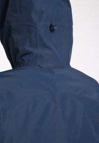 Vue arrière d'une personne portant une veste bleu foncé avec une capuche ajustable équipée d'un petit cordon réglable pour un ajustement serré.