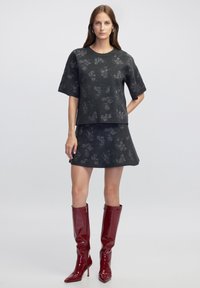 Diepgrijze, gestructureerde gebreide top met korte mouwen en bloemenpatroon, gecombineerd met een bijpassende skort. Model draagt glanzende rode knielange laarzen.