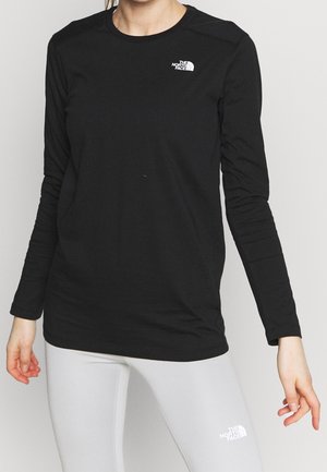 Kvinde iført en sort North Face langærmet trøje og hvide North Face leggings, stående med armene afslappet langs siden.