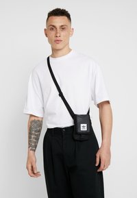 Bolsa de tela negra de bandolera con correa ajustable y parche de logo blanco. El modelo lleva una camiseta blanca holgada y pantalones negros, mostrando un brazo tatuado.