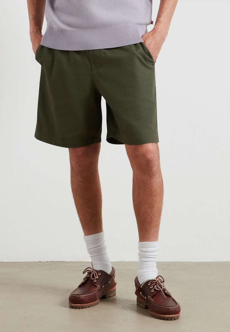 Shorts vert olive en tissu léger, avec des poches obliques et une coupe droite. Associés à des chaussures marron et des chaussettes blanches.