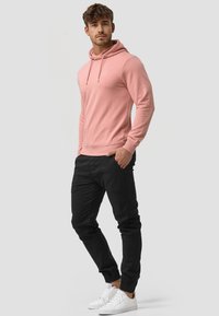 Svijetloroza hoodie s prednjim džepom i kapuljačom s vezicama, u kombinaciji s crnim tapered hlačama i bijelim tenisicama. Glatka tekstura i ležeran fit.
