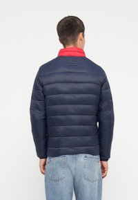 Marineblå pufferjakke med rød krave, vandret quiltet design og elastiske ærmer. Bæret over lyseblå denim, der har store baglommer.