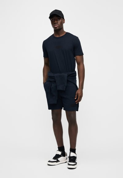 Tmavě námořnické tričko a šortky z bavlněné směsi, volný střih. Mikina s kapucí uvázaná kolem pasu, černá čepice a černo-bílé tenisky.