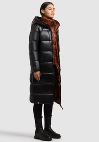 Kvinde iført en lang sort pufferjakke med et brunt indre lag, sorte leggings og sorte kraftige støvler, stående mod en ensfarvet lys baggrund.