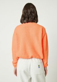 Oranžinis, pūkuotas megztinis su apvalia iškirpte, ilgomis rankovėmis ir laisvo kirpimo, derinamas su pilkomis sportinėmis kelnėmis, turinčiomis galinę kišenę.