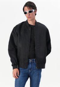 Chaqueta bomber negra con cremallera en la parte delantera, puños y dobladillo acanalados, combinada con una camiseta negra y jeans de mezclilla azul. Presenta gafas de sol blancas.