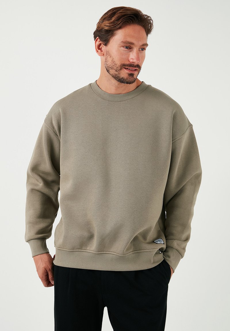 Sudadera verde olivo hecha de tela texturizada, con cuello redondo, hombros caídos y una pequeña etiqueta con el logo en el dobladillo.