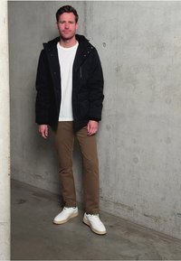 Svart zip-up jacka, vit t-shirt och bruna byxor. Skorna har vita sneakers med beige detaljer. Slät, strukturerad betongbakgrund.