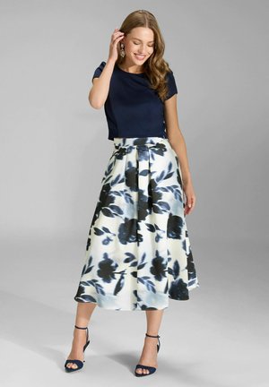 Kobieta ubrana w granatowy krótki rękaw, białą spódnicę midi z dużym czarnym kwiatowym wzorem oraz granatowe sandały na kostkę, uśmiecha się trzymając rękę przy włosach.