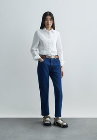 Bílá košile s dlouhým rukávem a knoflíkovou légou s nařasenými detaily, modré vysoké džíny, hnědý pásek a robustní sandály v béžové barvě s černými podrážkami.