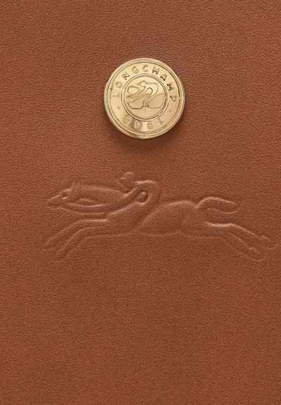 Goldener runder Knopf mit dem Schriftzug "LONGCHAMP 1948" darüber, geprägt auf Leder, das ein springendes Pferdelogo auf einer braunen strukturierten Oberfläche zeigt.